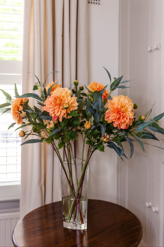 The Austell,  a faux floral arrangement, real-touch domed orange Dahlias with buds,  Eucalyptus Nicolii  foliage in a glass vase on a wooden table.