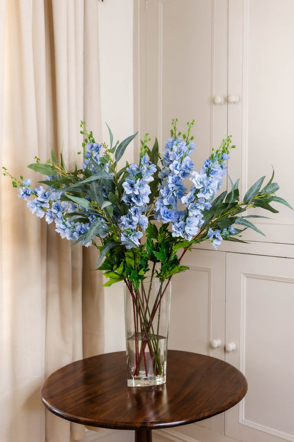 The Morden arrangement, seven faux real-touch blue Delphinium Larkspur stems, five stems of faux Eucalyptus Nicolii, in a glass vase on a wooden table.