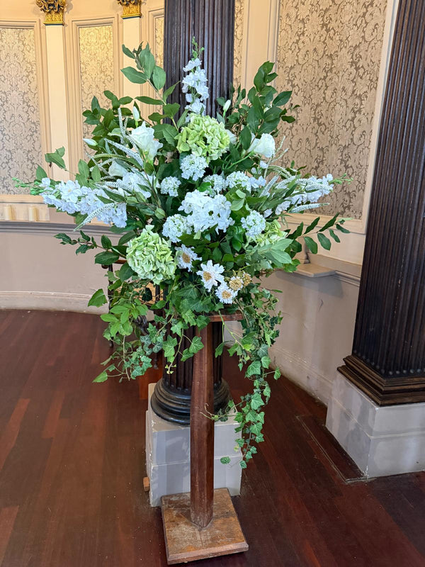 A large faux floral altar pedestal installation for the Church of the Ascension Blackheath, consisting of real touch green hydrangeas, white hydrangeas, white scabiosas,  ivory lisianthus, white delphiniums, white Veronica, white geraniums and lots of natural green foliage arranged in a bowl on top of an oak stand 