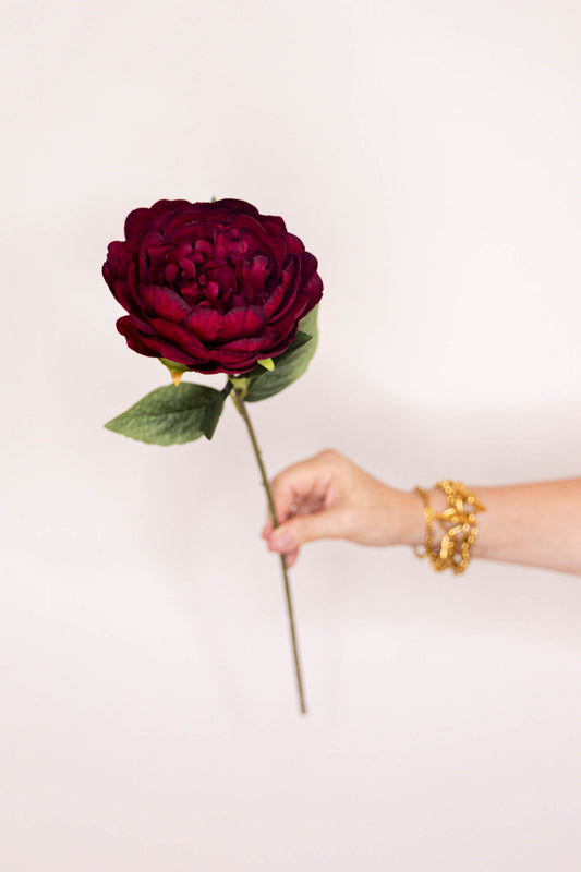 A single faux floral dark burgundy Peony  with green leaves on a long stem.