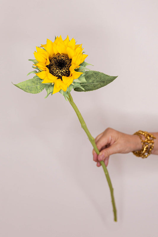A single artificial sunflower with bright yellow petals, a dark brown center, and green leaves.