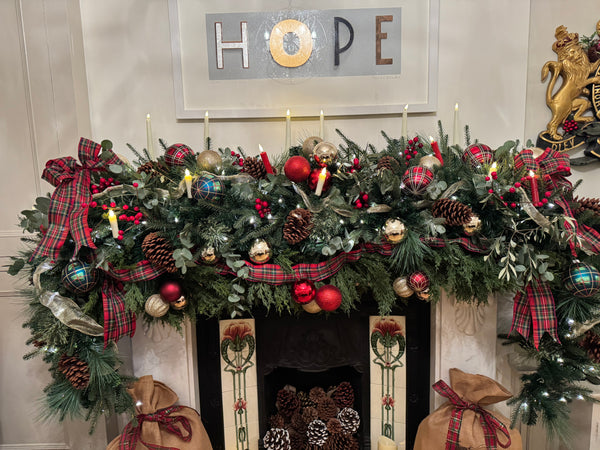 Faux Floral Christmas installation on a mantelpiece red and green tartan ribbon, tartan baubles, gold baubles, red baubles, candles, greenery, and fir cones.